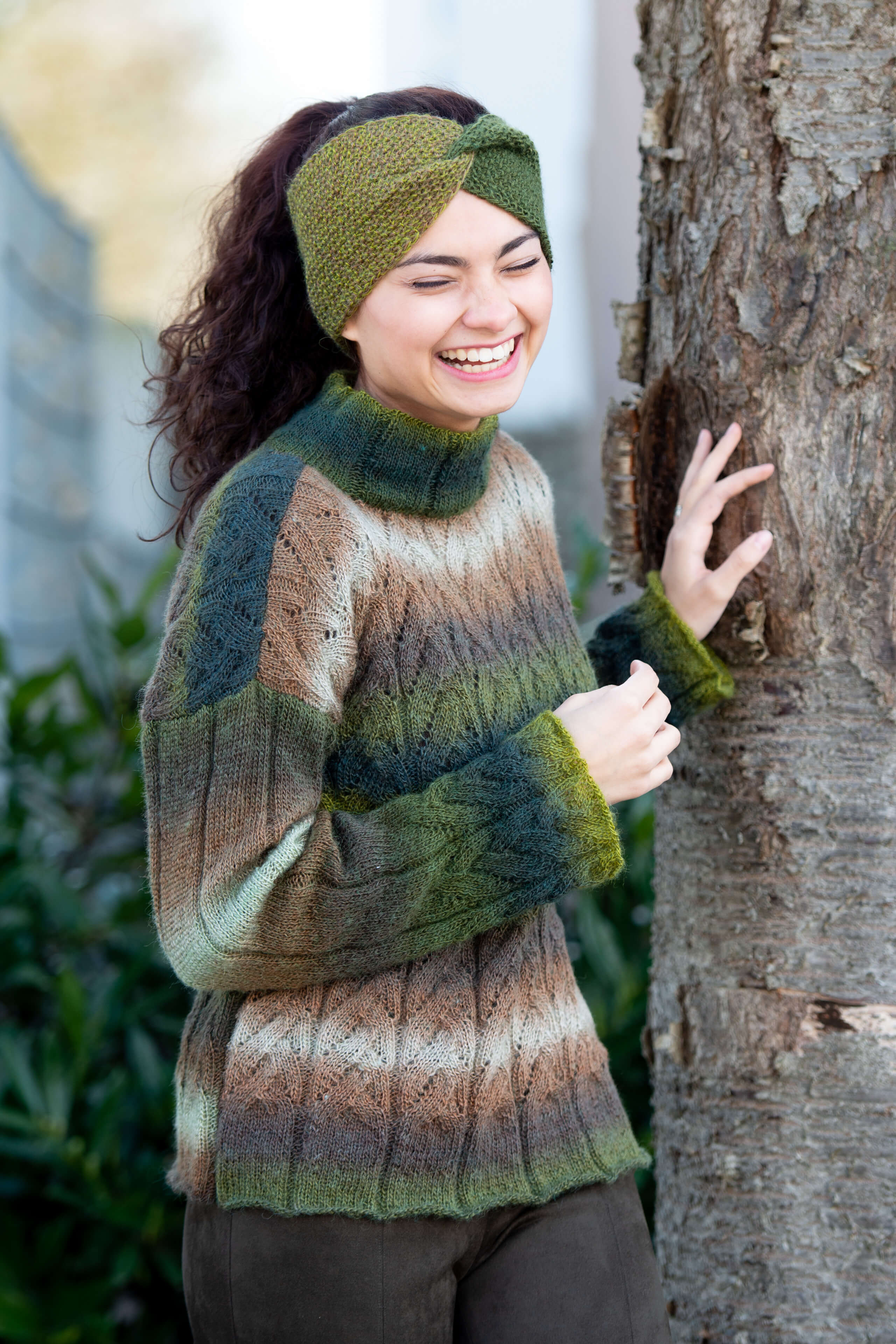lachende Frau abgestützt an Baum mit gestricktem grün-braunen Pullover und Stirnband