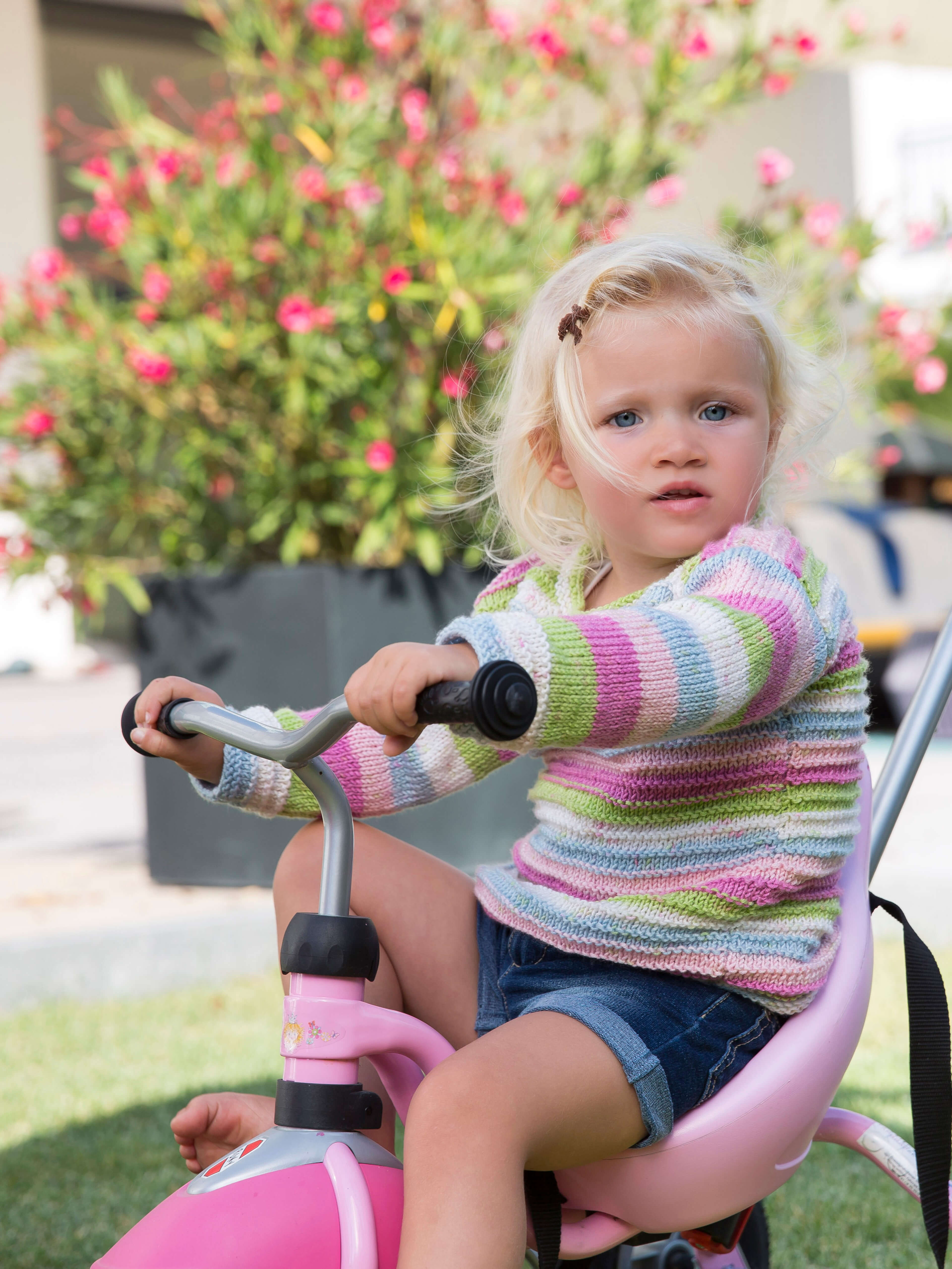 blondes Mädchen in grün-blau-rosa gestreiftem Pulli auf einem rosa Dreirad